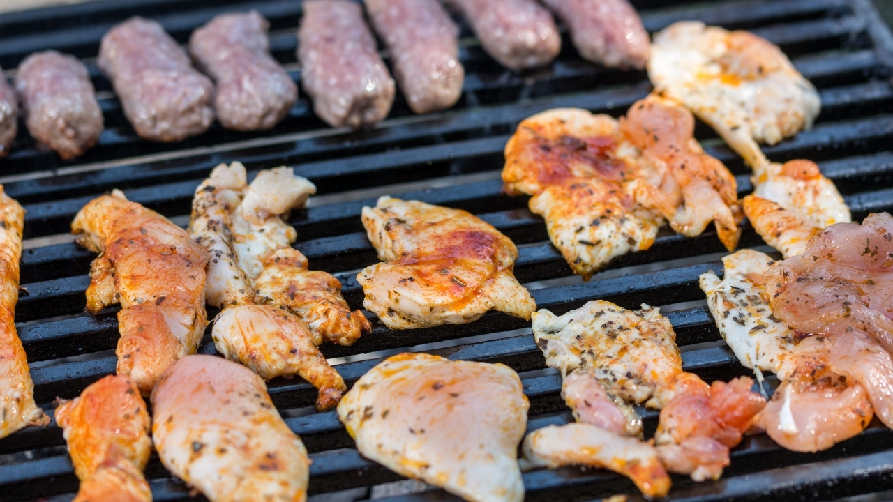 Komadi piletine i ćevapi raspoređeni na rešetki roštilja tokom pripreme